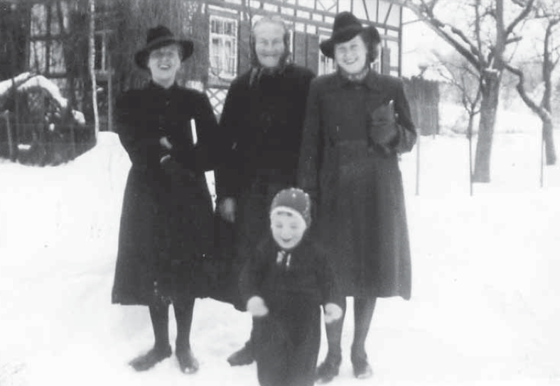 Historische Schwarz-Weiß-Fotografie einer winterlichen Szene. Drei Frauen in dunklen Mänteln und Hüten stehen lächelnd im Schnee vor einem Fachwerkhaus. Vor ihnen steht ein kleines Kind mit Mütze und Winterkleidung. Im Hintergrund sind schneebedeckte Bäume zu sehen.