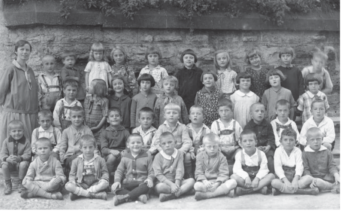 Historische Schwarz-Weiß-Gruppenfotografie einer Schulklasse mit zahlreichen Jungen und Mädchen unterschiedlichen Alters. Die Kinder sitzen und stehen in mehreren Reihen vor einer Steinmauer. Links steht eine erwachsene Frau, vermutlich die Lehrerin.