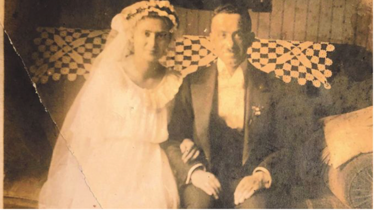 Historische, sepiafarbene Hochzeitsfotografie eines Paares. Die Braut trägt ein helles Hochzeitskleid mit Schleier und Blumenschmuck im Haar. Der Bräutigam sitzt neben ihr in einem dunklen Anzug. Im Hintergrund ist eine gemusterte Stoffdecke zu sehen.